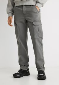 Άτομο που φοράει γκρι παντελόνι cargo με πλαϊνές τσέπες και μαύρα αθλητικά παπούτσια, στέκεται με το ένα χέρι στην τσέπη.