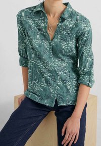 Camicia verde con bottoni, caratterizzata da un motivo nautico, maniche arrotolate e un fit rilassato. Indossata con pantaloni in denim scuro, che mettono in risalto le texture.