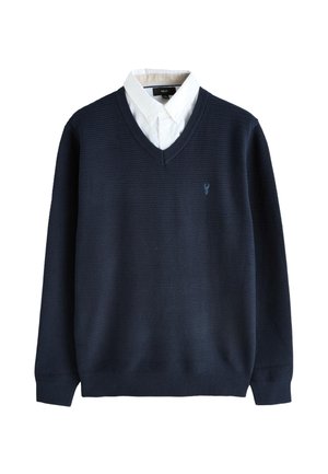 Granatowy sweter z dekoltem w serek o fakturowanym splocie, noszony na białej koszuli zapinanej na guziki, z małym haftowanym logo na lewej piersi.