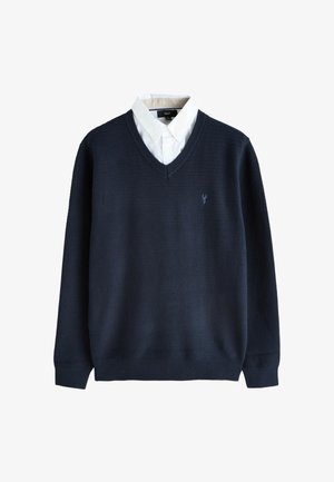 Granatowy sweter z dekoltem w serek o fakturowanym splocie, noszony na białej koszuli zapinanej na guziki, z małym haftowanym logo na lewej piersi.