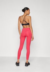 Črni športni modrček z nazaj prekrižanimi naramnicami, v kombinaciji z živahnimi rožnatimi leginami z visokim pasom. Črne športne čevlje dopolnjujejo videz.