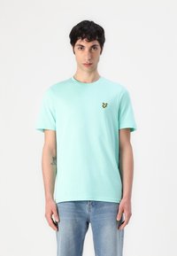 T-shirt en coton vert menthe avec un col rond, des manches courtes et un logo de papillon jaune sur la poitrine gauche. Porté avec un jean bleu clair.