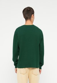 Donkergroene langemouwenshirt van een textuurstof, met een ronde halslijn en een ontspannen pasvorm. Zichtbaar stikselontwerp op de mouwen.