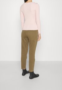 Donna in piedi di spalle, con una blusa a maniche lunghe rosa chiaro, pantaloni verde oliva e stivaletti neri, su uno sfondo bianco semplice.