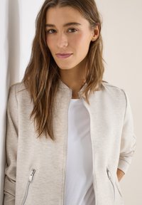 Jeune femme aux longs cheveux bruns portant une veste zippée beige clair et une chemise blanche, debout devant un fond neutre.