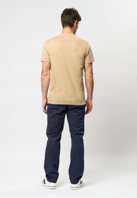 Beige kortermet t-shirt med rund hals, parret med marineblå bukser og hvide sneakers, set fra ryggen.
