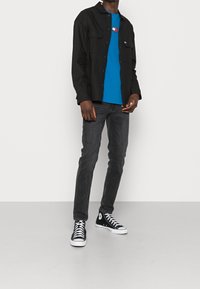 Czarna koszula z długim rękawem na niebieskiej koszulce z logo, w połączeniu z ciemnymi dżinsami i czarnymi trampkami za kostkę. Gładkie tło.