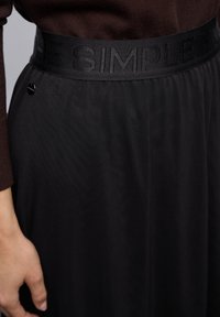 Jupe noire avec une taille texturée ornée du texte en relief "THE SIMPLE". Un petit détail circulaire noir est visible près de la taille.