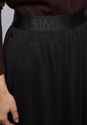 Zwarte rok met een gestructureerde tailleband met daarop het reliëf "THE SIMPLE" tekst. Een klein zwart, rond detail is zichtbaar nabij de tailleband.