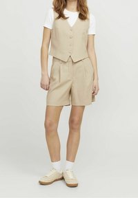 Beige shorts with pleats paired with a matching vest featuring three buttons, worn over a white T-shirt. Footwear: beige sneakers with light accents.