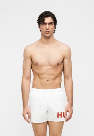 Homme musclé aux cheveux bouclés foncés portant un short de bain blanc avec les lettres rouges « HU », debout devant un fond clair uni.