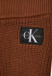 La tela de punto acanalada marrón presenta una etiqueta negra con "Calvin Klein Jeans" y un logotipo blanco de "CK" exhibido de forma prominente.