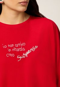 Femme portant un haut rouge avec un texte italien brodé : "io non arrivo in ritardo creo Suspenso" décoré de sequins sur "Suspenso".
