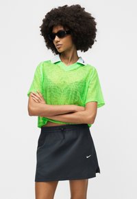 Γυναίκα με αφροκάν μαλλιά, φοράει μαύρα γυαλιά ηλίου, διάφανο τοπ με νεον πράσινο μοτίβο και λευκό γιακά, καθώς και μαύρη φούστα Nike, σταυρωμένα χέρια.