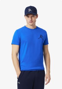 Sininen puuvillapaita, jossa on pyöreä pääntie ja lyhyet hihat, ja vasemmassa rinnassa musta siluettilogot. Yhdistetty tummansinisiin housuihin ja lippikseen.