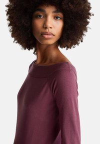 Mladá žena s kudrnatými vlasy v mauve svetru s odhalenými rameny, která hledí přímo před sebe s neutrálním výrazem.