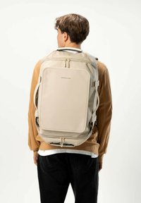 Sac à dos beige au design structuré, avec une texture lisse, des fermetures à double zip et des bretelles réglables. Logo de la marque affiché en évidence.