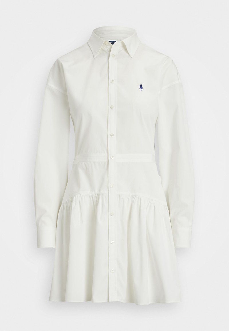 Bela srajčna obleka z dolgimi rokavi, nagubanim krilom in prilega pasu. Na prsih je majhna mornarsko modra oznaka.