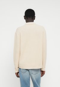 Μπεζ πουκάμισο με μακριά μ�ανίκια, γιακά και ριμπ μανσέτες, με διακριτικό ζιγκ-ζαγκ σχέδιο, συνδυασμένο με ανοιχτόχρωμο τζιν παντελόνι.