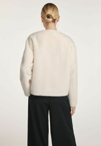 Kobieta z blond włosami w kok posiadająca puszysty, biały sweter z długim rękawem oraz szerokie czarne spodnie, stojąca tyłem do aparatu na jednolitym tle.