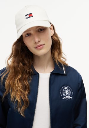 Noor naine pikkade laineliste juustega, seljas valge Tommy Jeans'i nokamüts, tumesinine embleemiga jakk ja valge särk, taustaks ühevärviline taust.