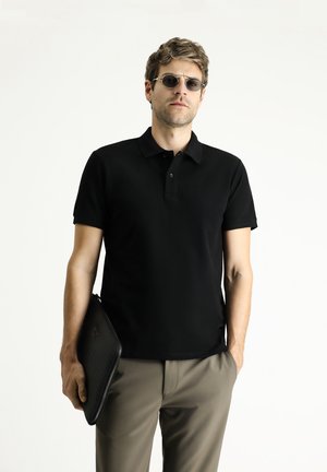 Camisa polo negra con cuello clásico y mangas cortas, combinada con pantalones beige. El modelo sostiene un bolso de mano negro texturizado.