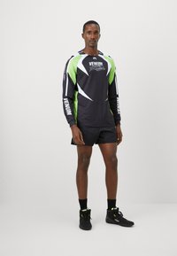 Schwarzes Sportshirt mit langen Ärmeln und grünen sowie weißen Akzenten, ausgestattet mit einem grafischen Logo und perforierten Details, kombiniert mit schwarzen Shorts und Schuhen.