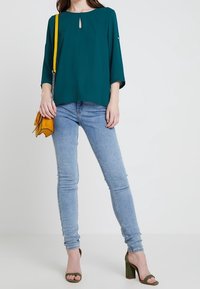 Turkusowa bluzka z detalem w kształcie dziurki, z rękawami do trzech czwartych, zestawiona z jasnoniebieskimi rurkami i oliwkowymi sandałami na obcasie. W zestawie żółta torebka.