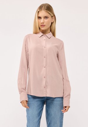 Donna che indossa una camicia rosa chiaro a righe verticali con bottoni e jeans blu, in piedi davanti a uno sfondo semplice.