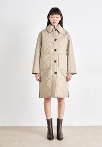 Manteau matelassé beige avec un col, doté de grands boutons, de poches latérales et d'une coupe jusqu'aux genoux. Porté par-dessus des bottes à talons marron.