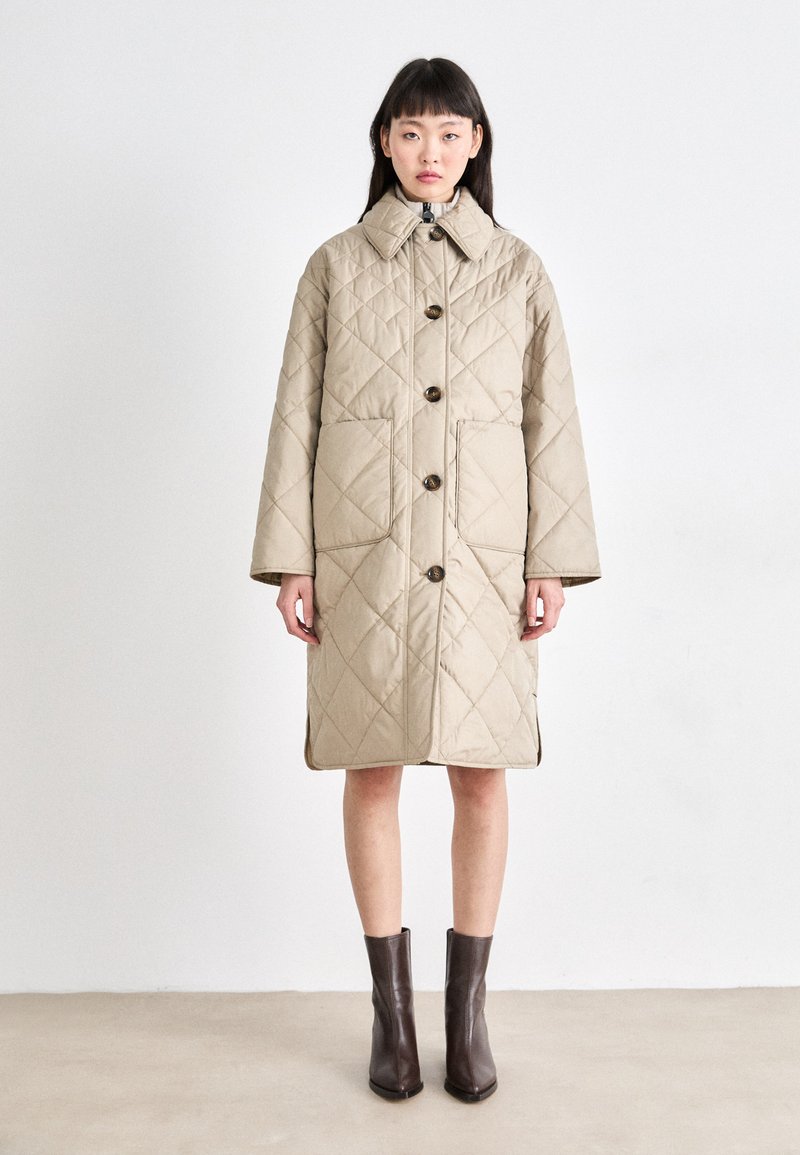 Manteau matelassé beige avec un col, doté de grands boutons, de poches latérales et d'une coupe jusqu'aux genoux. Porté par-dessus des bottes à talons marron.