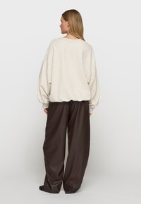 Cremefarbener Oversized-Pullover mit fallenden Schultern, gerippten Bündchen und lockerem Sitz, kombiniert mit weiten braunen Hosen aus glattem Stoff.
