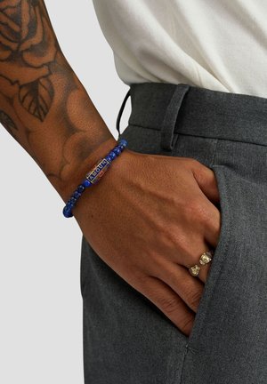 Blaues Perlenarmband mit einem goldenen Mittelstück, das das Wort „AMOUR“ zeigt, getragen an einer Hand mit Ringen, vor grauer Hose.