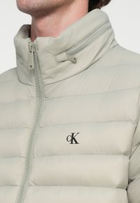 Casaco acolchoado cinza claro com design matelassê, gola alta e fecho de correr; apresenta um logótipo preto "CK" no peito.