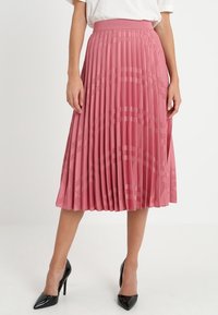 Falda midi plisada en rosa suave, con un acabado texturizado y una amplia cinturilla elástica. Combinada con tacones negros puntiagudos.