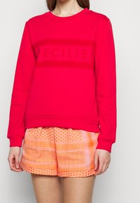 Röd sweatshirt med ribbad textur och "CECILIE"-logotyp över bröstet. Matchas med mönstrade orange shorts med geometriska mönster.