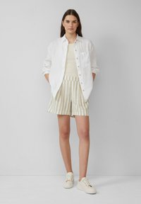 Ein weißes Leinenhemd über einem cremfarbenen Top, kombiniert mit beigen und weißen gestreiften Shorts. Das Model trägt hellfarbene Sneakers und steht vor einem neutralen Hintergrund.