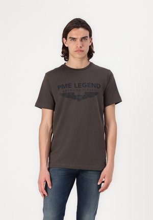 T-shirt marron foncé en coton doux, avec un imprimé graphique noir bien visible et des manches courtes. Associe à un jean bleu.