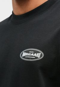 Schwarzes Baumwoll-T-Shirt mit einem gerippten Rundhalsausschnitt und einem weißen ovalen Logo mit "MADS NØRGAARD COPENHAGEN" auf der Vorderseite.