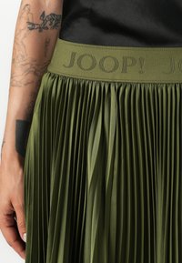 Jupe plissée vert olive avec une large taille ornée du logo "JOOP!". Fabriquée en tissu lisse, mettant en avant des plis verticaux et une texture douce.