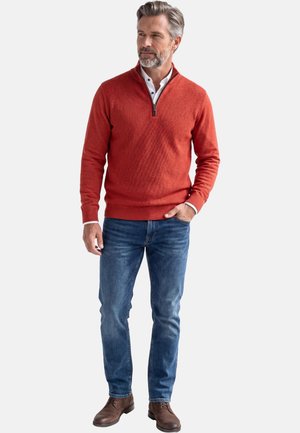 Mann mit grauem Haar und Bart, der einen orangefarbenen Pullover mit Reißverschluss, ein weißes Hemd, blaue Jeans und braune Lederschuhe trägt, mit einer Hand in der Tasche stehend.