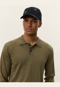 Olijfgroen gebreid poloshirt met een kraag en drie zwarte knopen, gecombineerd met een zwarte pet met driehoekige grijze accenten.