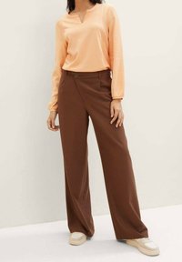 Langarm, gerippte orange Bluse mit einem leichten V-Ausschnitt. Kombiniert mit hochtaillierten, weiten braunen Hosen und cremefarbenen Sneakers.