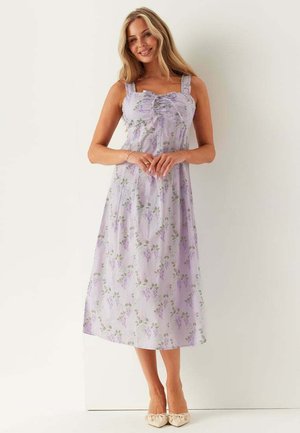 Kvinde står iført en ærmeløs lavendel midi-kjole med blomsterprint og beige spidse flade sko, smilende mod en ensfarvet baggrund.