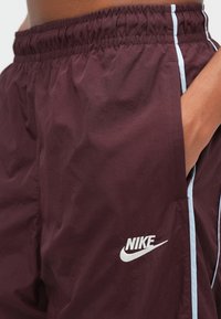 Bordové šortky Nike s elastickým pásom, bočnými vreckami a svetlomodrými bočnými pruhmi. Na prednej strane je biele logo Nike.