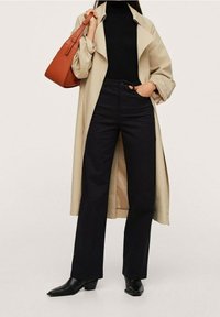 Manteau long beige sur un col roulé noir, associé à un jean noir taille haute et des bottines noires brillantes. Sac à main structuré marron.