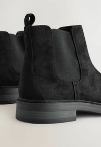 Botines de ante negros con paneles elásticos en los laterales y un tacón bajo, posicionados sobre un fondo claro, destacando la textura y el diseño.