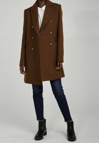 Hnědý dvoj řadý vlněný kabát se zlatými knoflíky, klopami a bočními kapsami. Stylizováno přes bílou košili a tmavé džíny s černými kotníkovými botami.