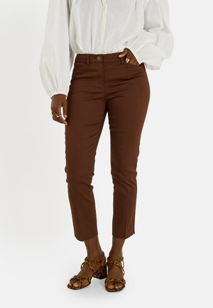 Persona con pantalones marrones ajustados, blusa blanca de manga larga y sandalias de tacón con estampado de leopardo, de pie con una mano en el bolsillo.