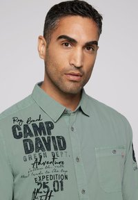Lichtgroen overhemdresshirt met geprinte logo's en tekst, een borstzak en een kraag. De stof lijkt lichtgewicht en gestructureerd.